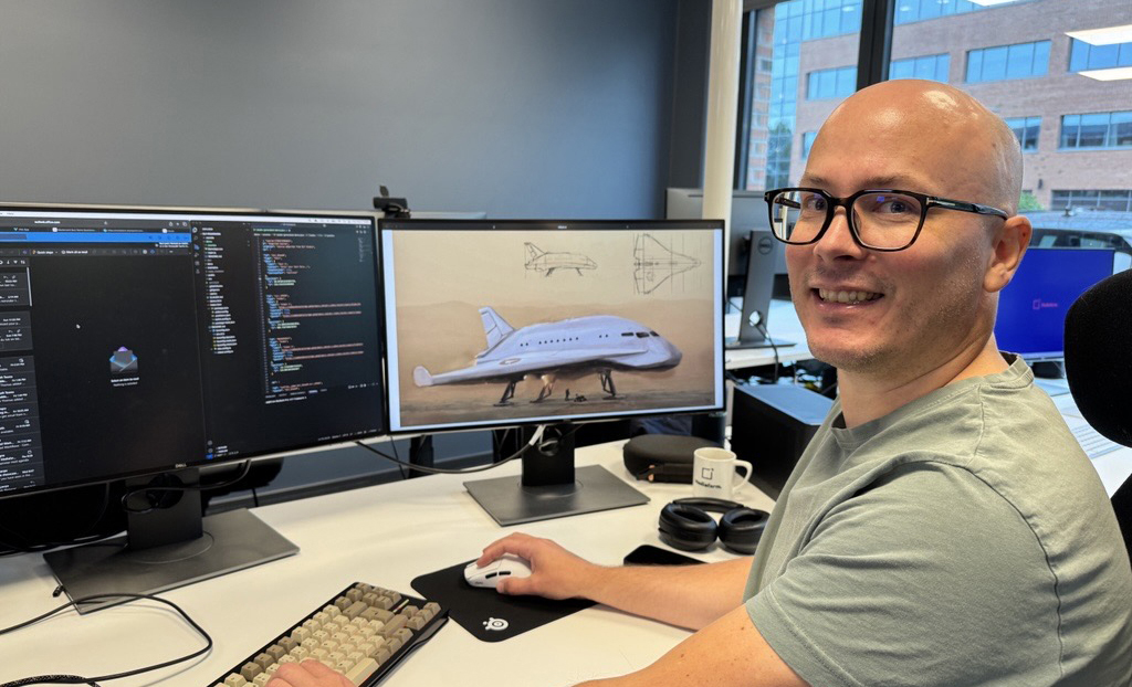 En mann med briller sitter foran to pc skjermer og smiler.