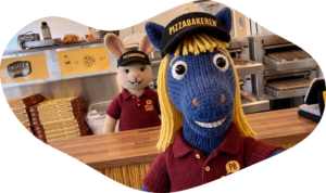 En lilla, strikket dukkehest har på seg Pizzabakercaps og t-skjorte og tar en selfie på et kjøkken. Bak hesten står en stikket kanin også iført uniform fra Pizzabakeren.