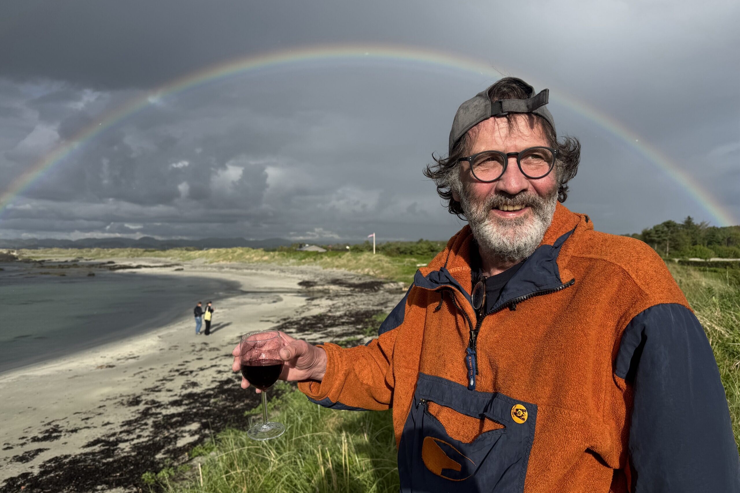Vilhelm st&aring;r foran en regnbue p&aring; Rege. Han smiler og holder et glass r&oslash;dvin i h&aring;nden.