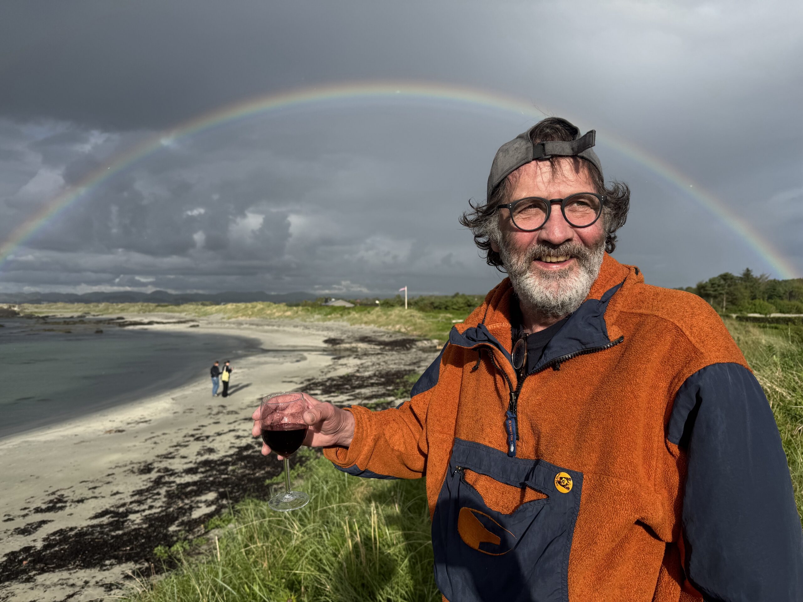 Vilhelm står foran en regnbue på Rege. Han smiler og holder et glass rødvin i hånden.